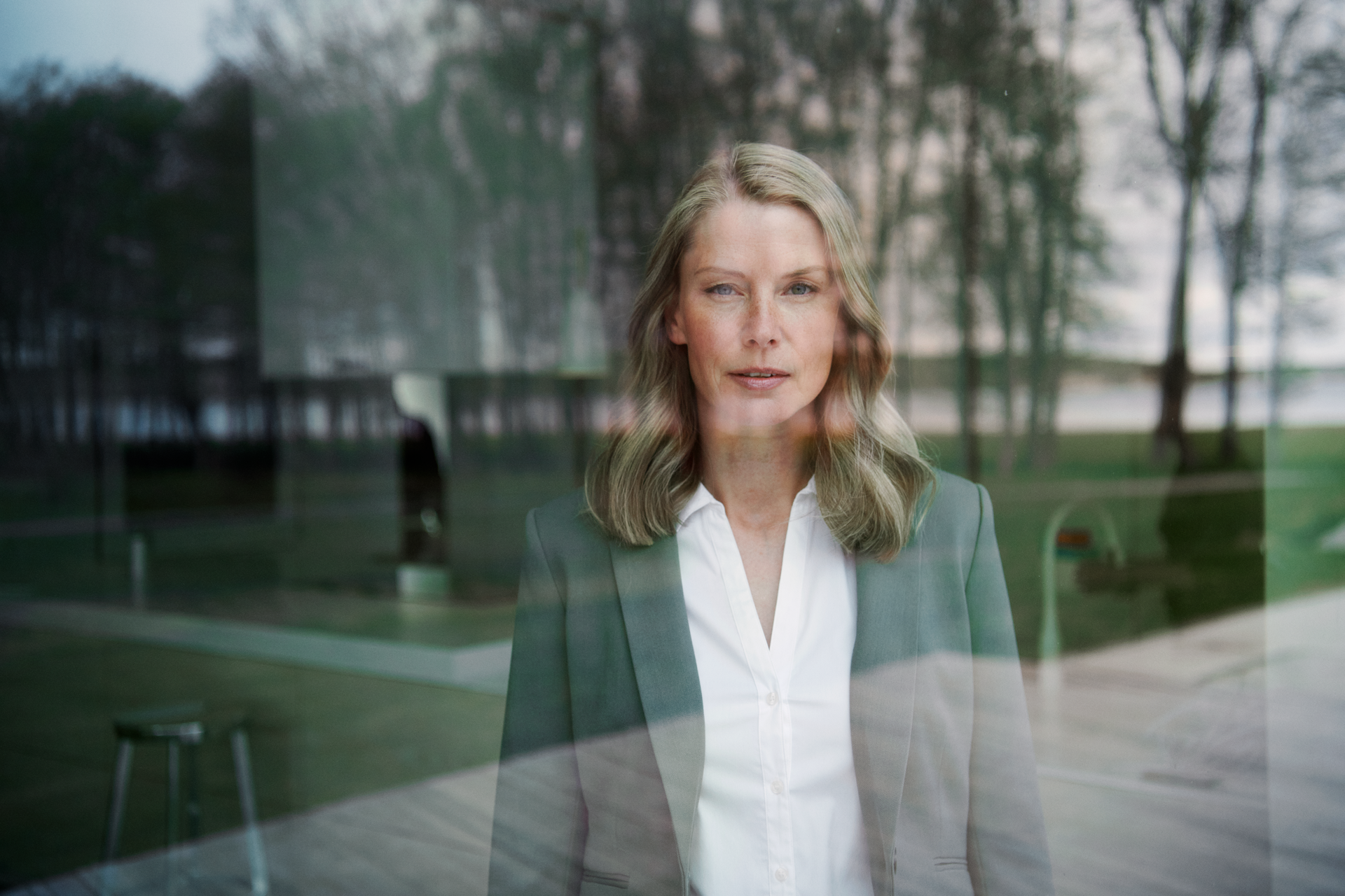 Vrouw met witte blouse en groen jasje bij raam met uitzicht op bomen - weet wat je niet moet doen bij energie besparen