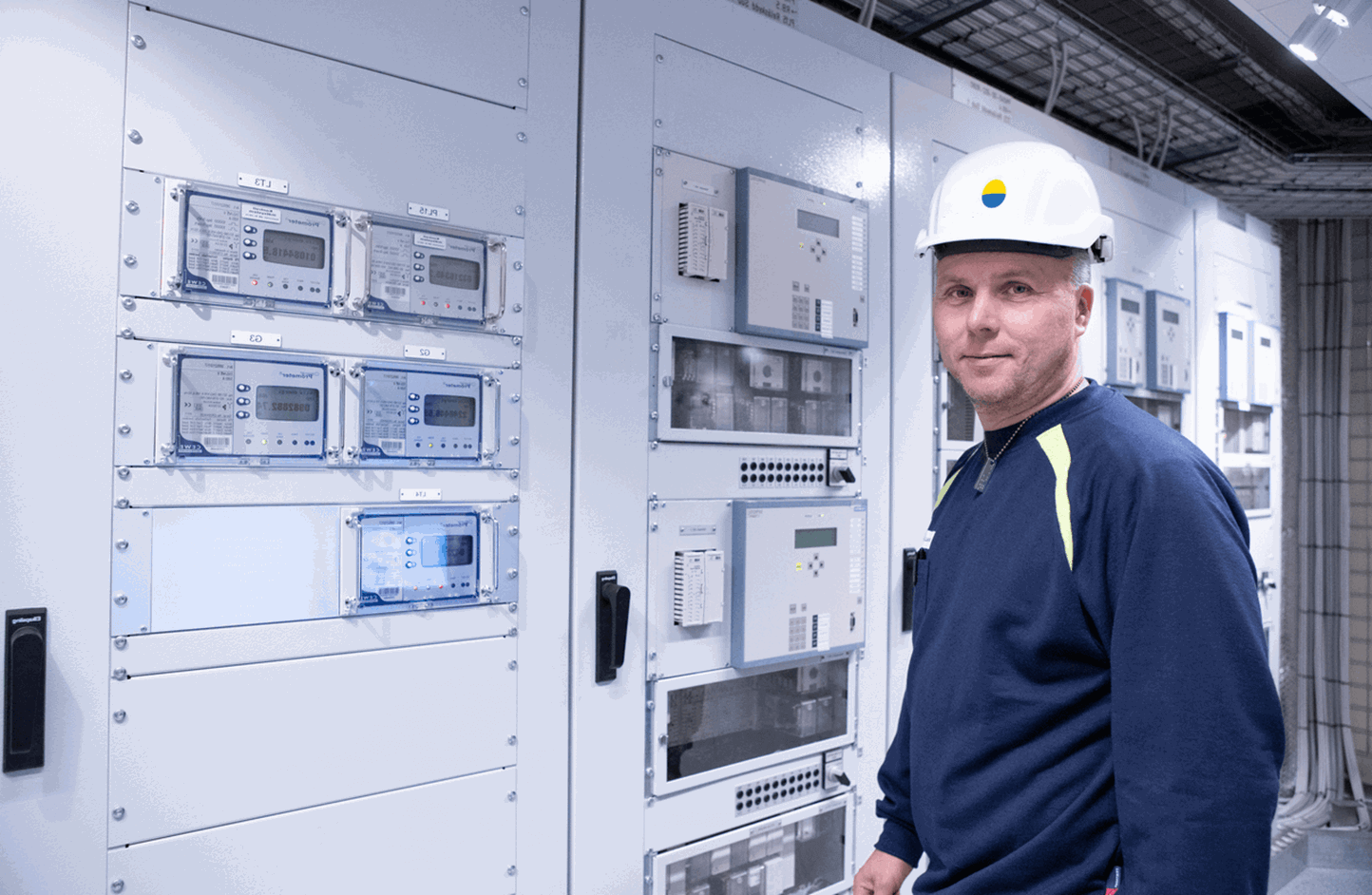 Man met helm die je aankijkt, met op de achterkant een technische installatie