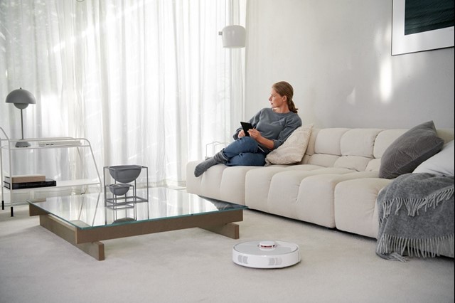 Vrouw zit op de bank in de woonkamer en de robot stofzuiger staat aan